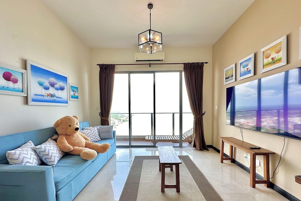 Una sala de estar con un ventanal grande, un sofá azul, un oso de peluche, un televisor y obras de arte en las paredes.