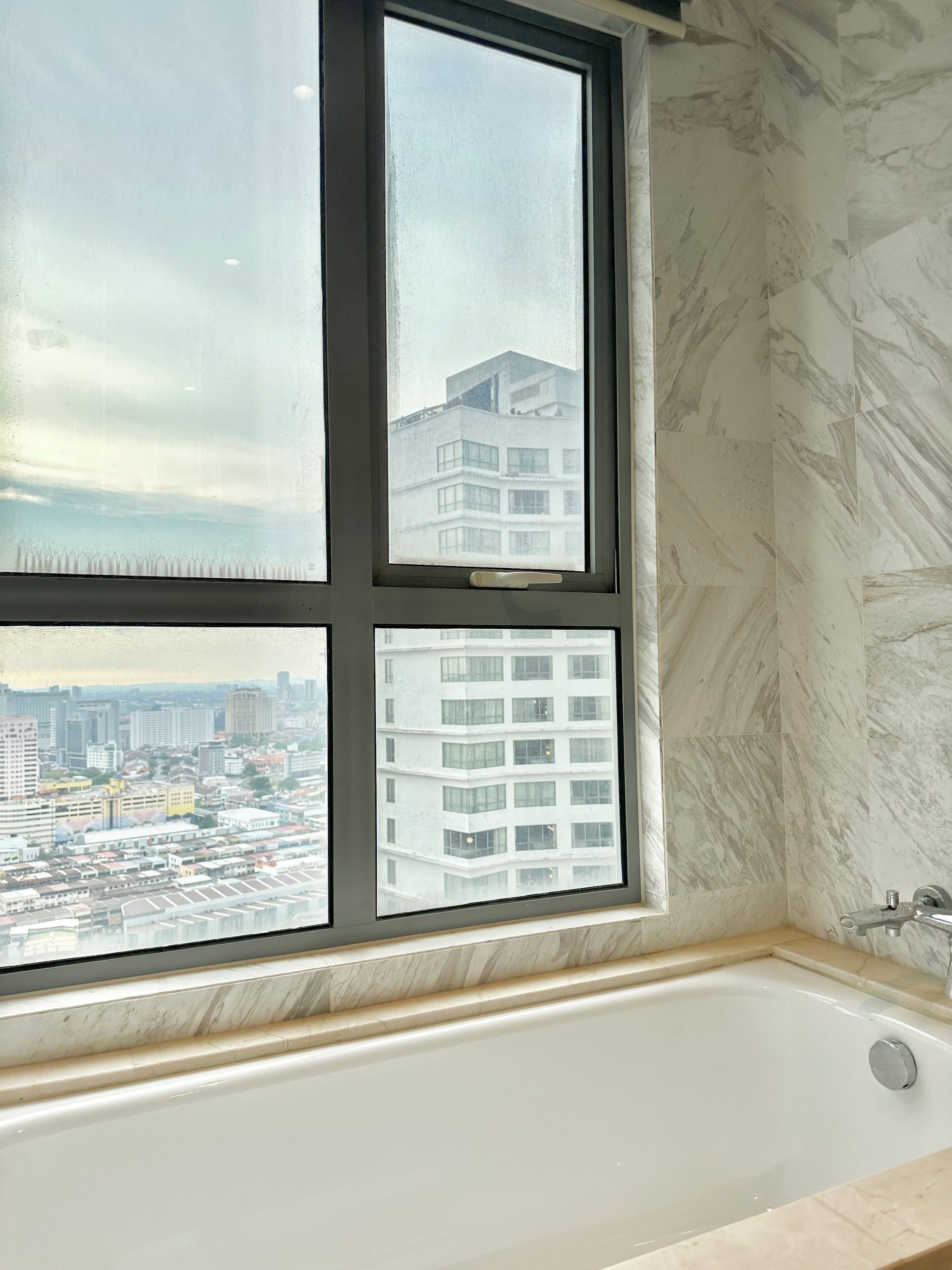 A bathroom with a bathtub, a window with a city view, and marble walls.