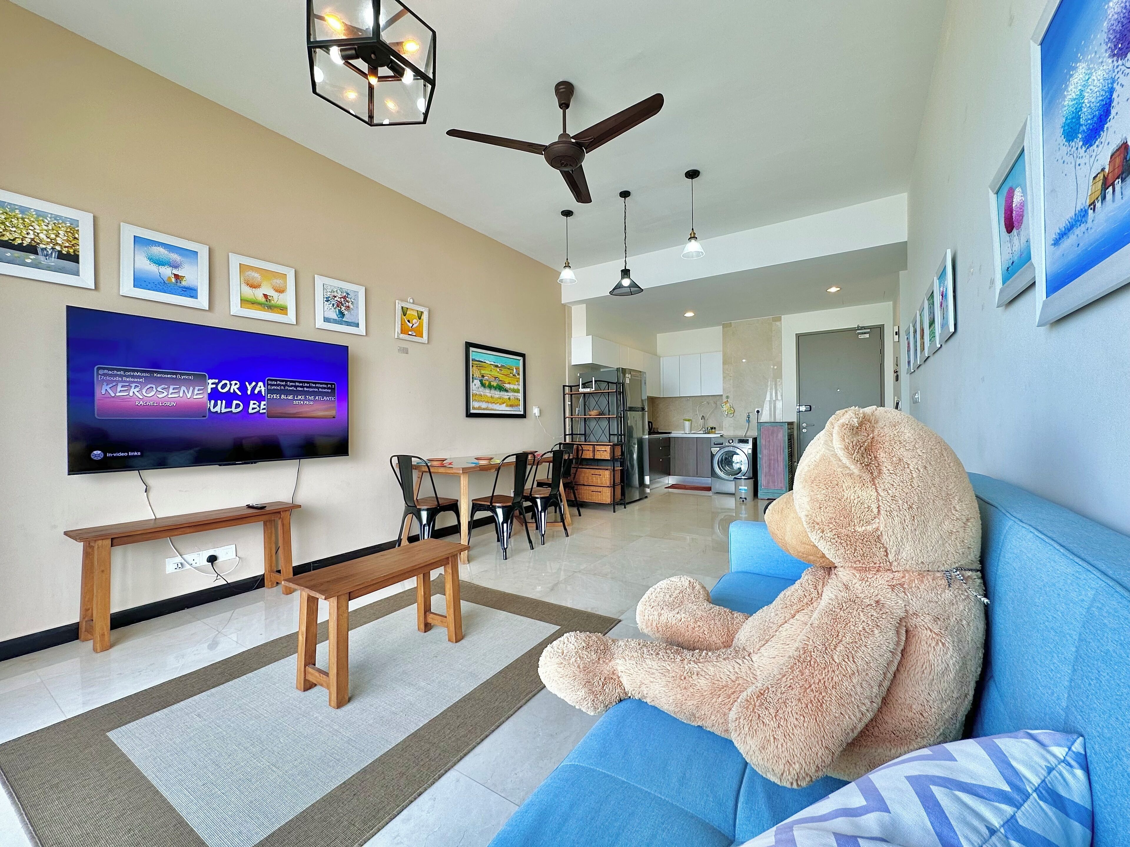 A living room with a large flat-screen TV, a ceiling fan, a dining area with a table and chairs, and a blue sofa with a teddy bear.