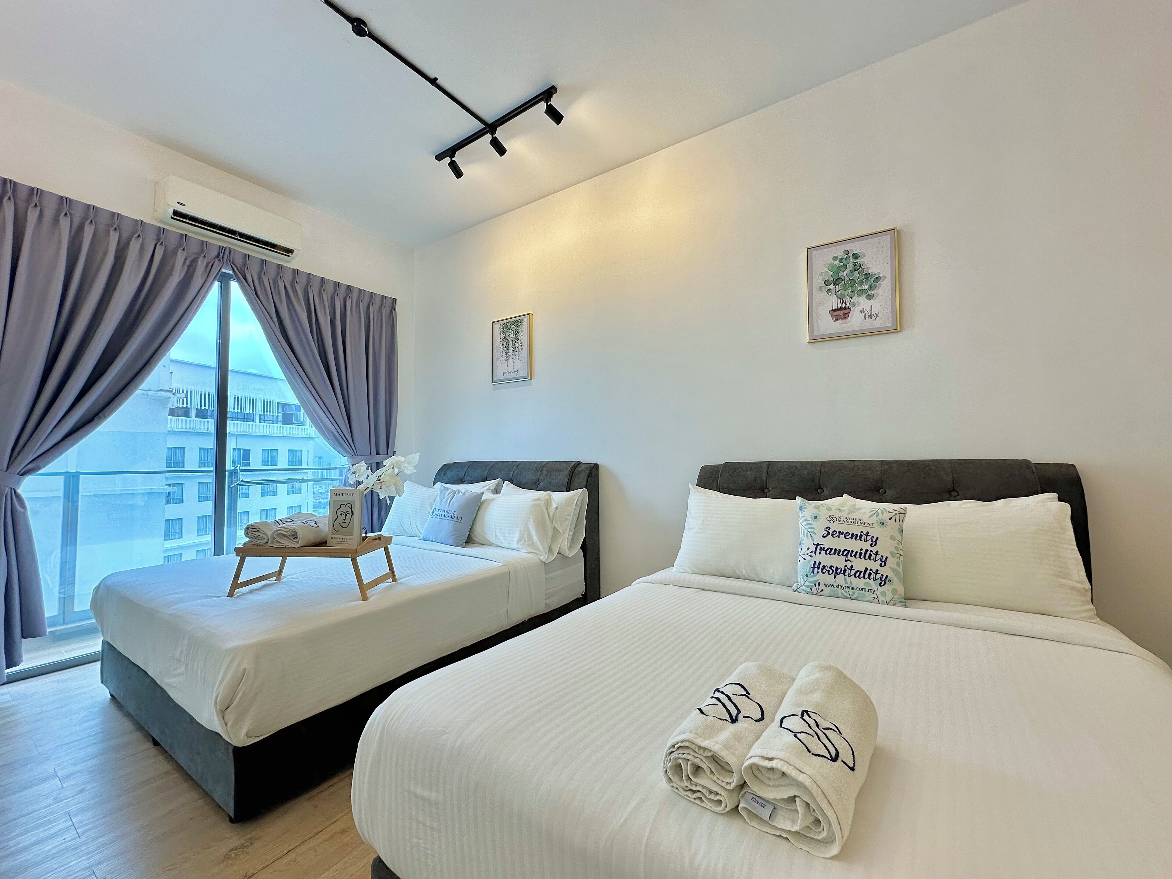 A hotel room with two beds, a large window with curtains, a ceiling-mounted air conditioner, and a small table with towels.