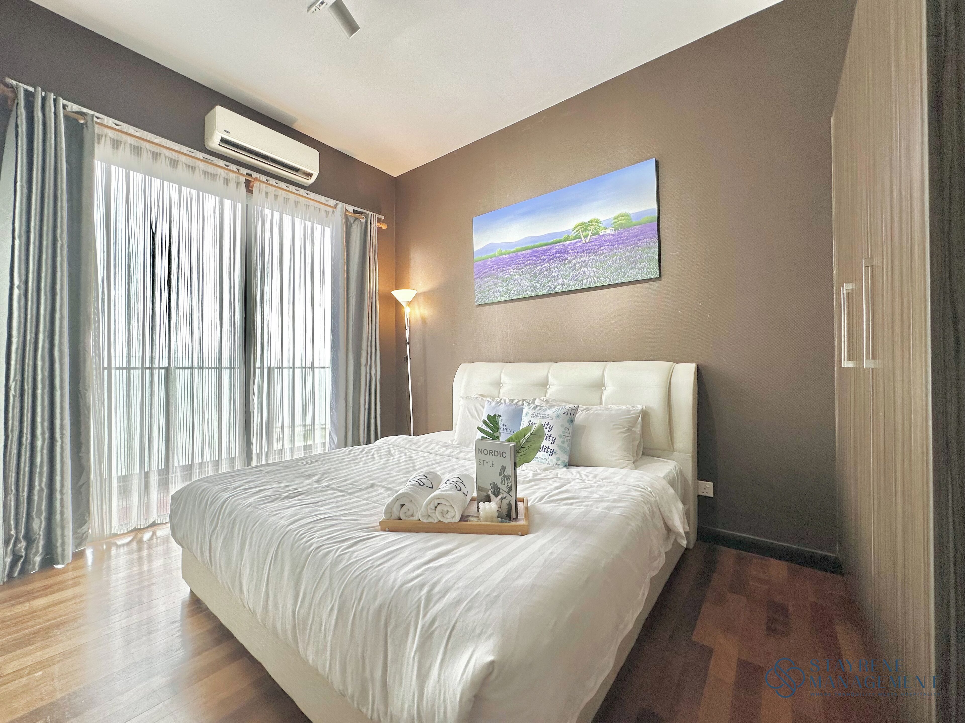 A hotel room with a large bed, a wall-mounted air conditioner, a painting of a landscape, and a bedside lamp.