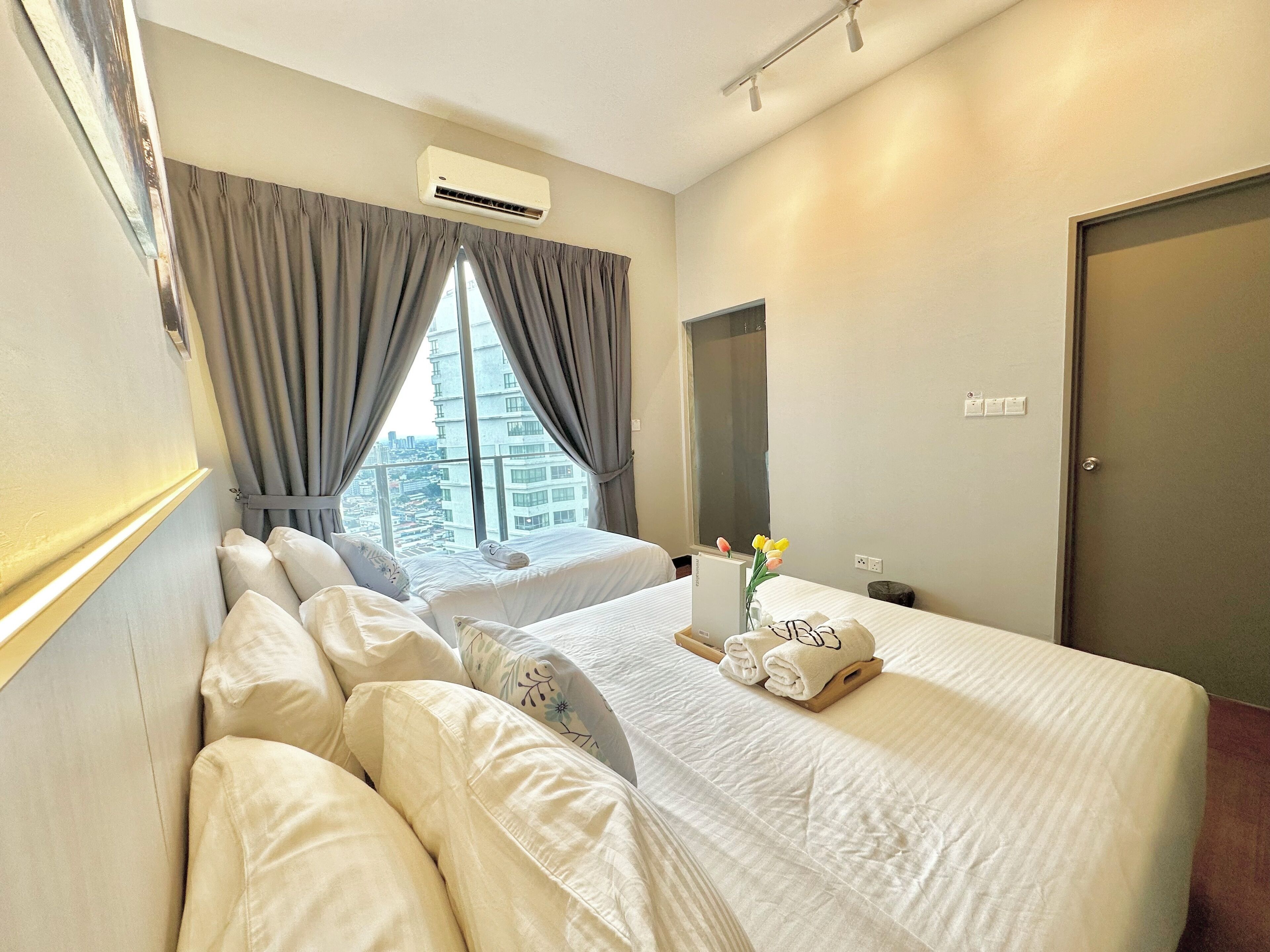 A hotel room with two beds, a window with curtains, an air conditioning unit, and a bedside table with a vase of flowers.