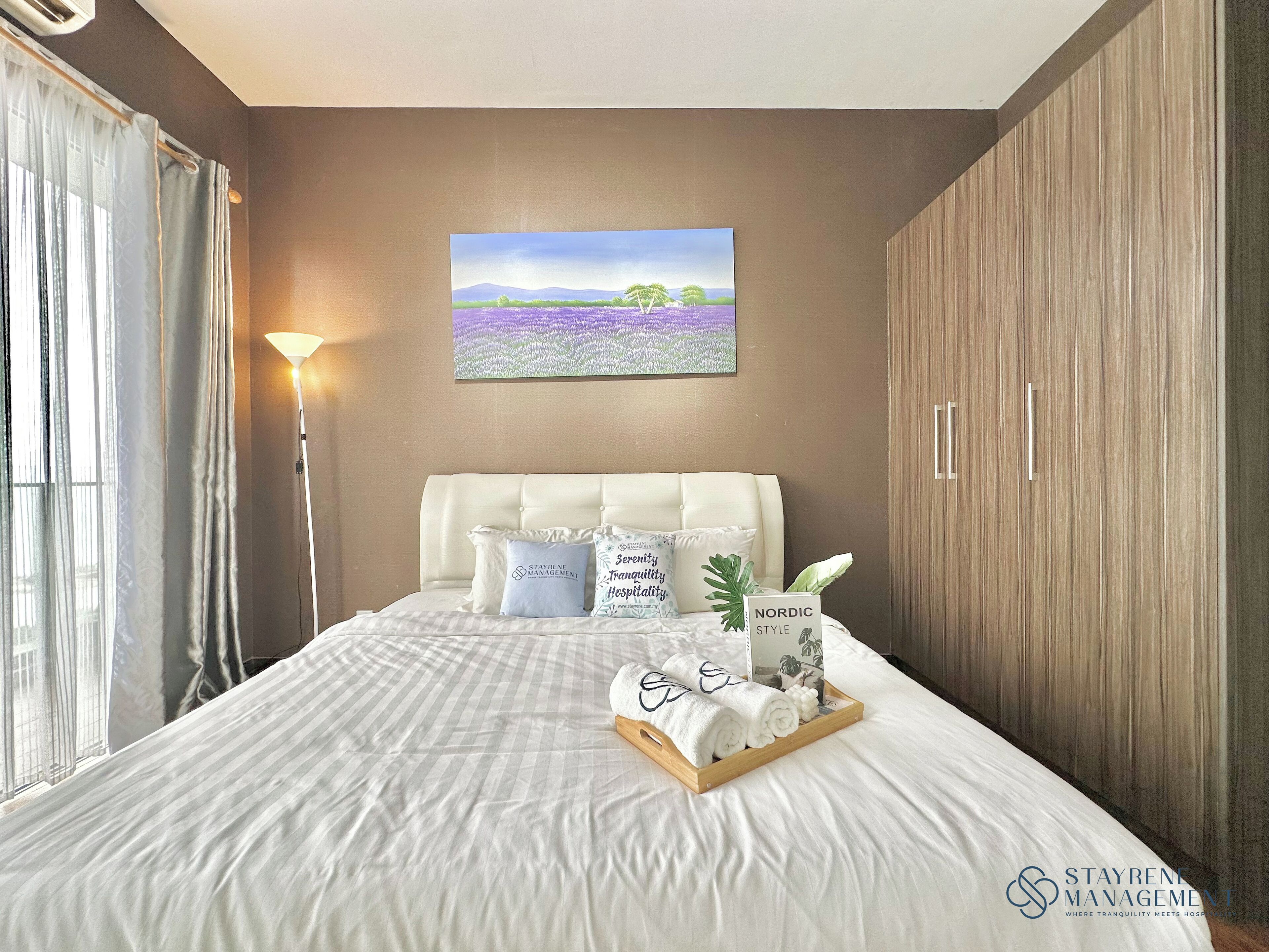 A hotel room with a large bed, a headboard, a bedside table with a tray of towels, a lamp, and a painting of a landscape.