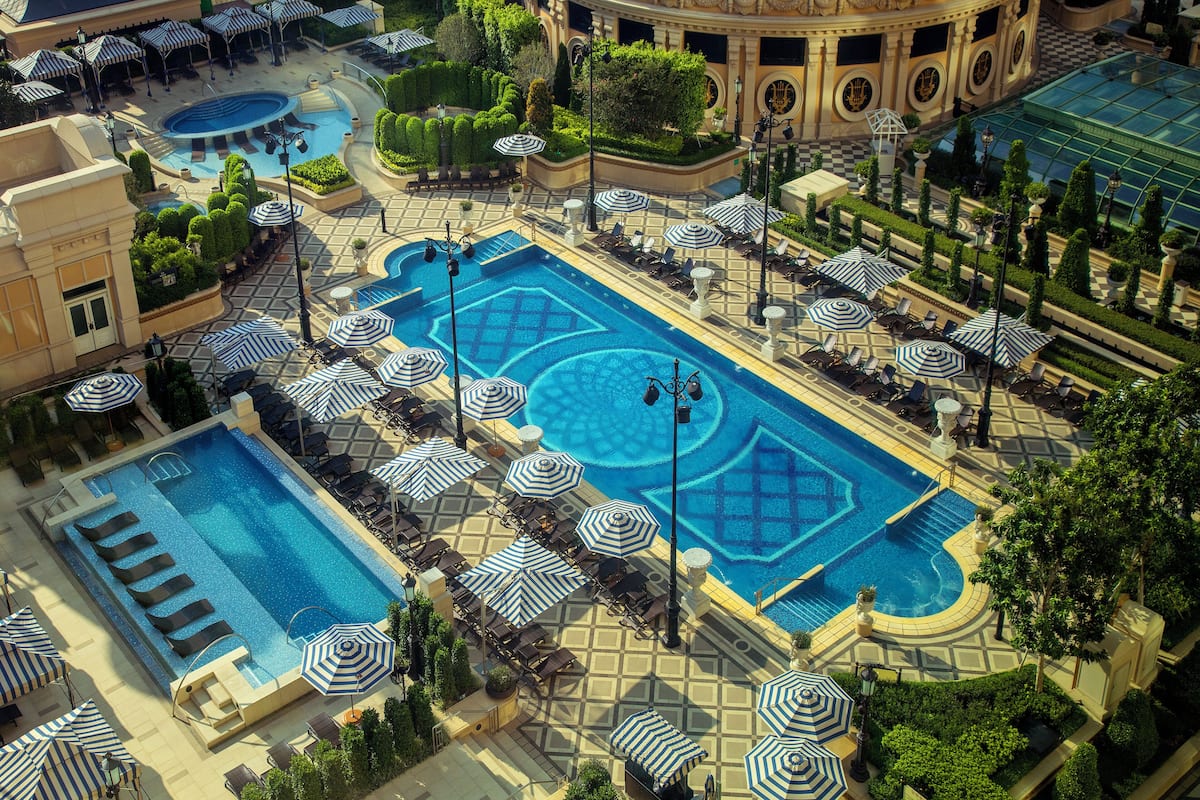 A large pool area with multiple lounge chairs and umbrellas, surrounded by well-manicured gardens and a grand building.