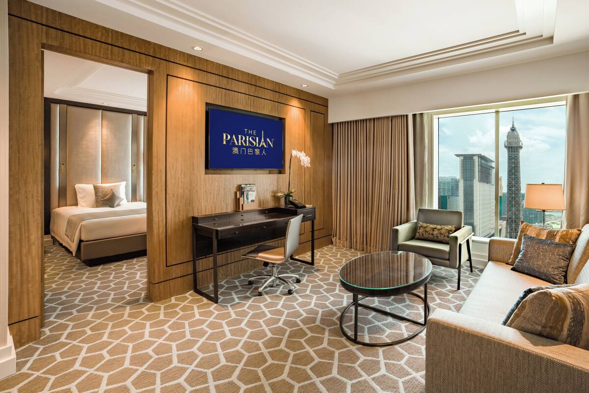 A hotel room with a bed, desk, chair, and sofa, a city view, and a sign reading 'The Parisian'.