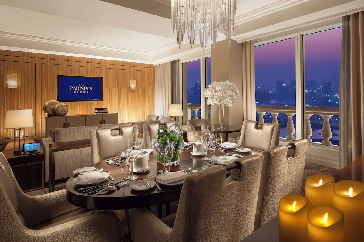 A dining area with a view of the city at dusk, a television displaying 'The Parisian,' and a chandelier.