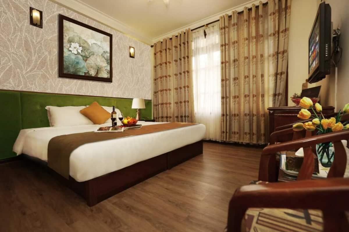 A hotel room with a bed, a nightstand, a lamp, a painting, curtains, a television, and a chair with a vase of flowers.