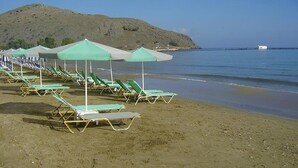 Ein Strand mit Reihen von Liegestühlen und Sonnenschirmen, ein sandiger Uferstreifen und eine Hügelkette im Hintergrund.
