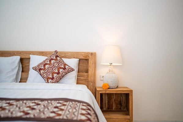 Ein ordentlich bezogenes Bett mit gemusterten Kissen, einem Holz-Kopfbrett und einem Nachttisch mit Lampe und einer dekorativen orangefarbenen Blüte.