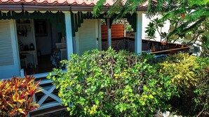 A house with a red tiled roof, a porch with a green railing, and a variety of plants in the foreground.
