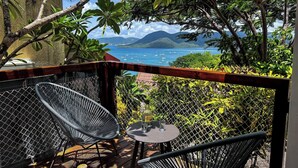 A balcony with wicker chairs and a small table, overlooking a scenic view of the sea and mountains.