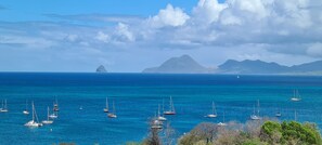 Un paysage côtier avec de nombreux voiliers sur une eau calme, un ciel dégagé et des montagnes à l’horizon.