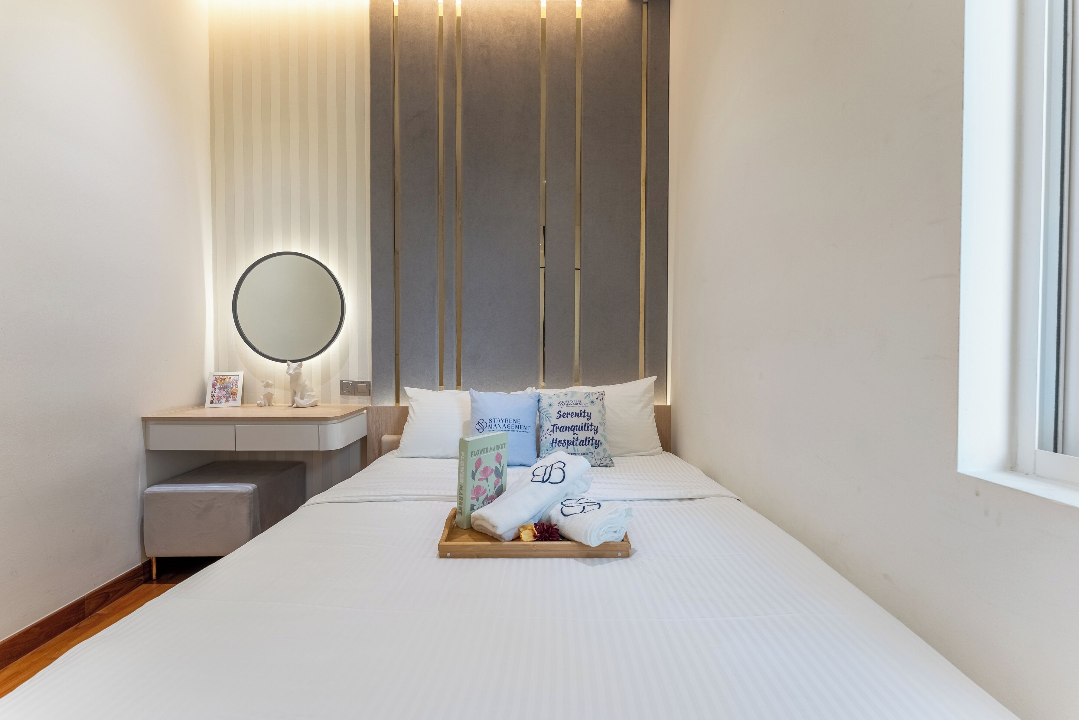 A neatly made bed with pillows, a bedside table with a mirror, a tray with towels and a book, and a window with blinds.