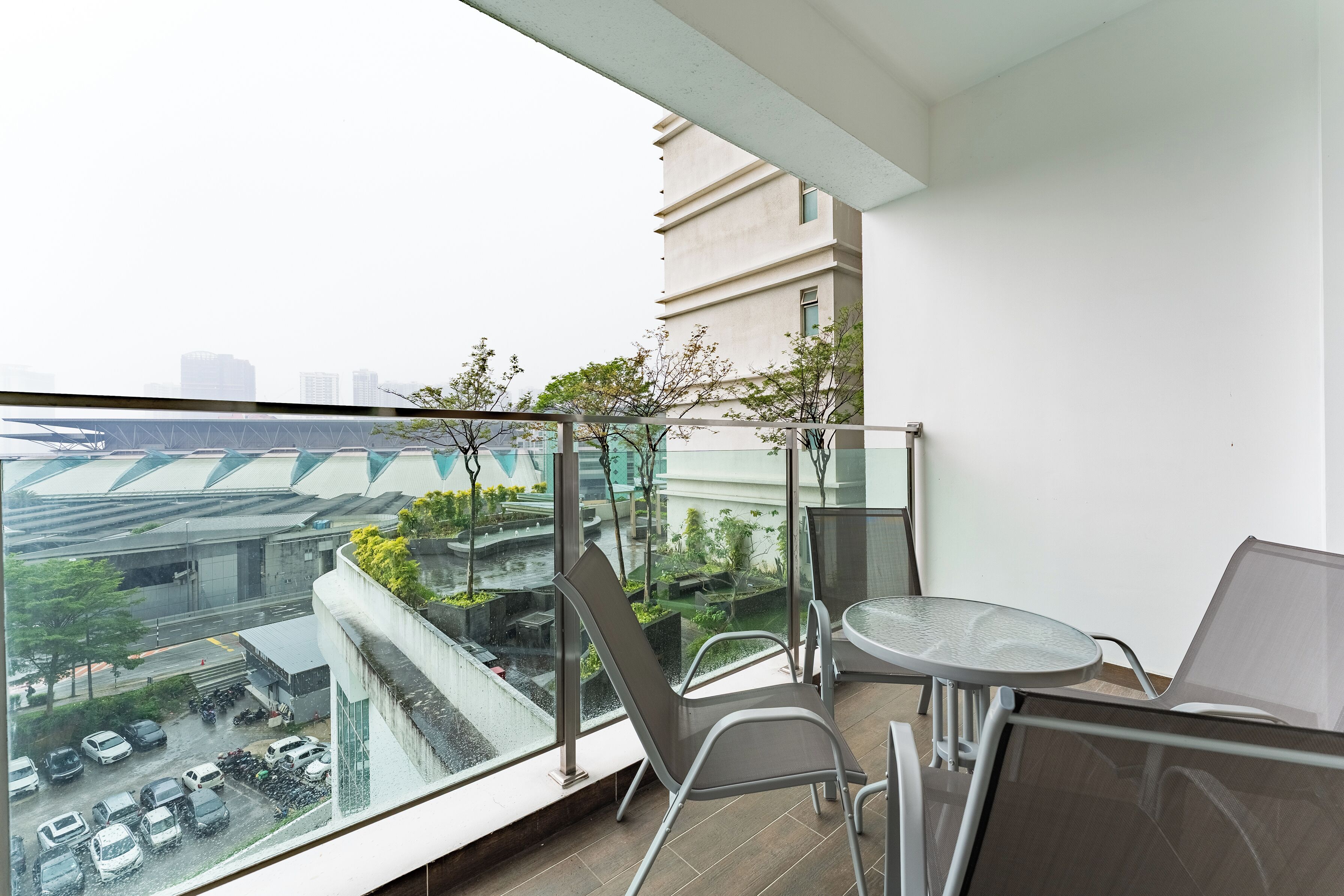 A balcony with a glass railing, a table, and chairs, overlooking a cityscape with buildings and a road.