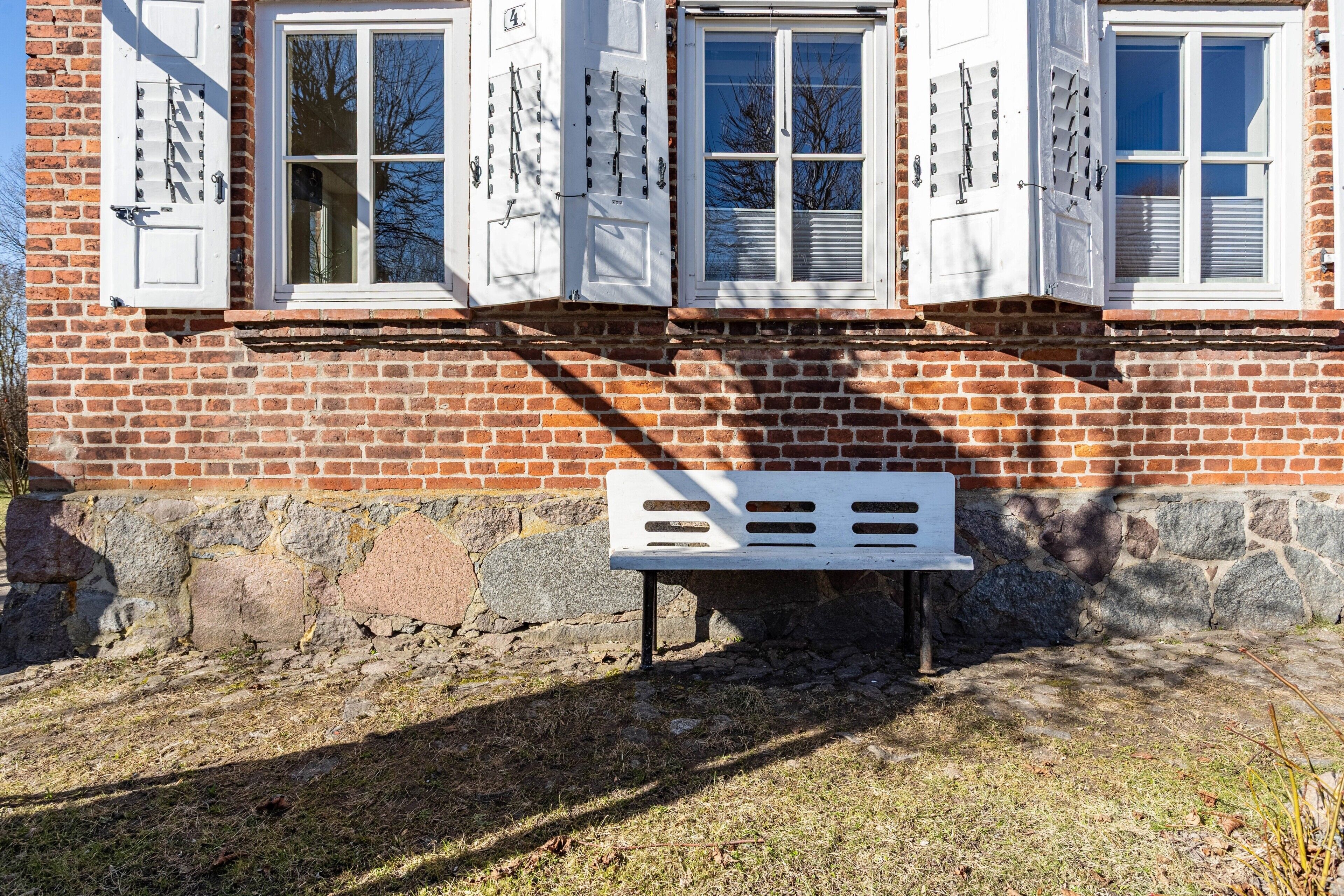 A brick building with three white-framed windows and shutters, a stone foundation, and a white bench in front.