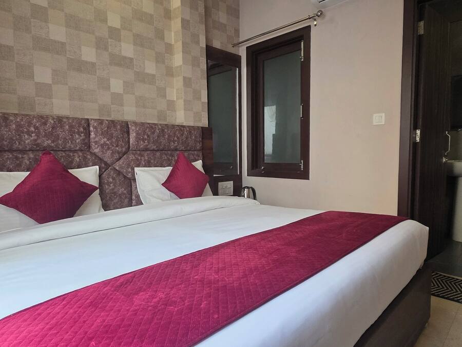 Una habitación de hotel con una cama grande, un cabecero de madera, dos almohadas rojas, una colcha blanca, una manta roja, una ventana, un baño con lavabo y espejo, y un patrón de pared a cuadros.