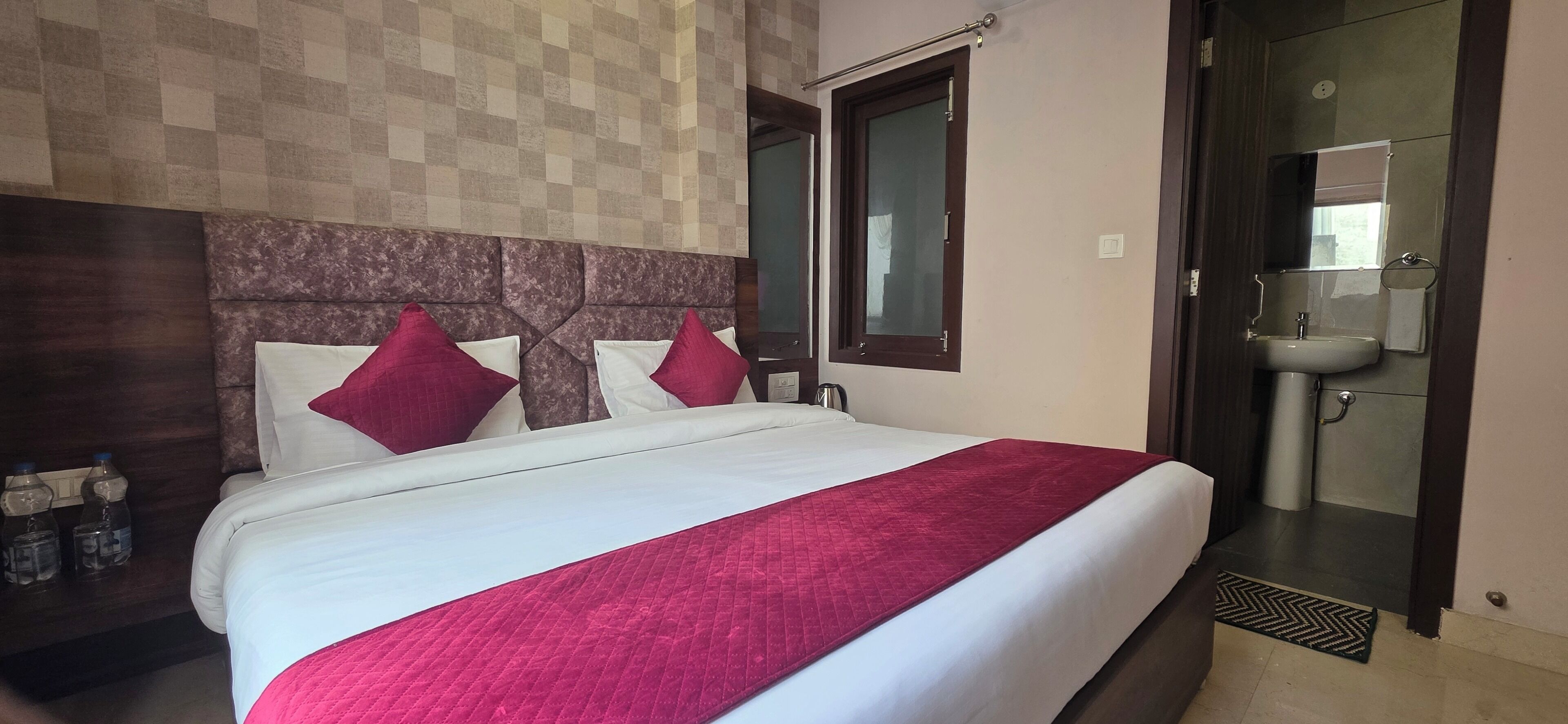 Une chambre d’hôtel avec un grand lit, une tête de lit en bois, une fenêtre, une salle de bain avec un lavabo et un miroir, et une porte.