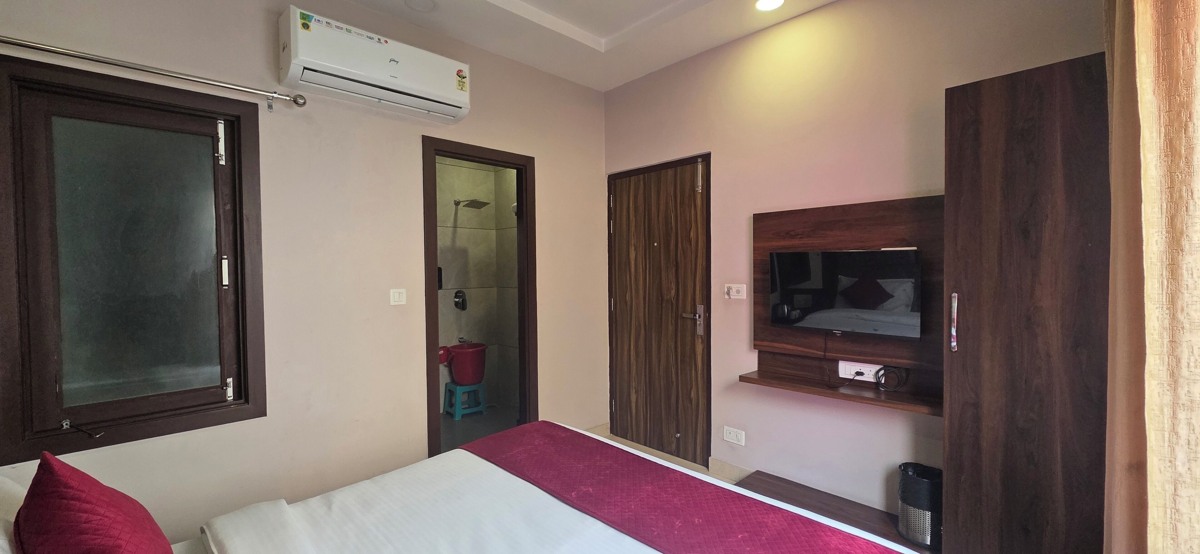 A hotel room with a bed, a TV mounted on the wall, a wooden wardrobe, and an air conditioning unit.