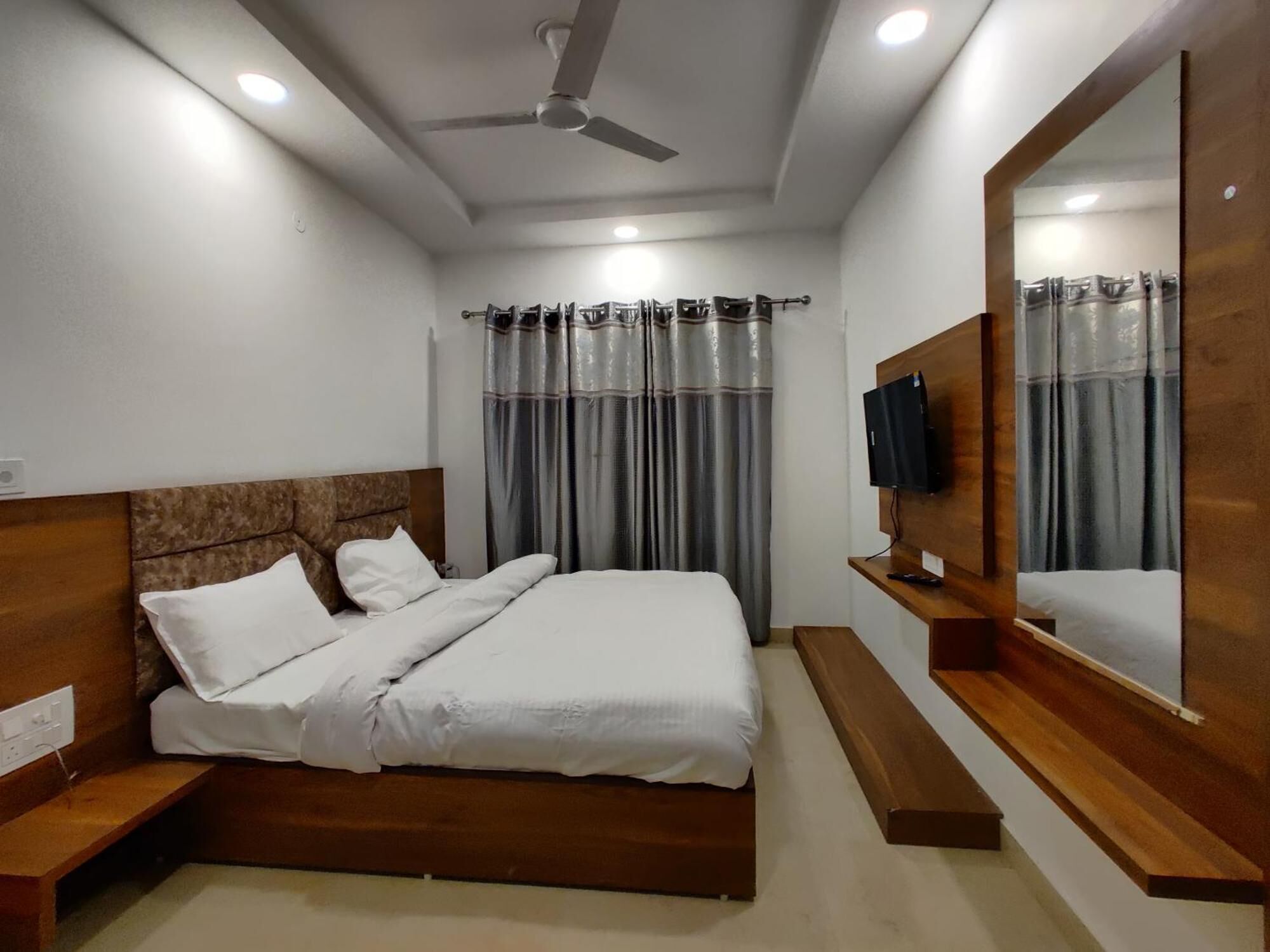A modern hotel room with a wooden headboard, a large mirror, a flat-screen TV, and a ceiling fan.