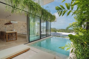 Un espace piscine extérieur moderne doté d’un patio fermé par des baies vitrées, d’un coin repas avec une table et des chaises, et d’une végétation luxuriante.