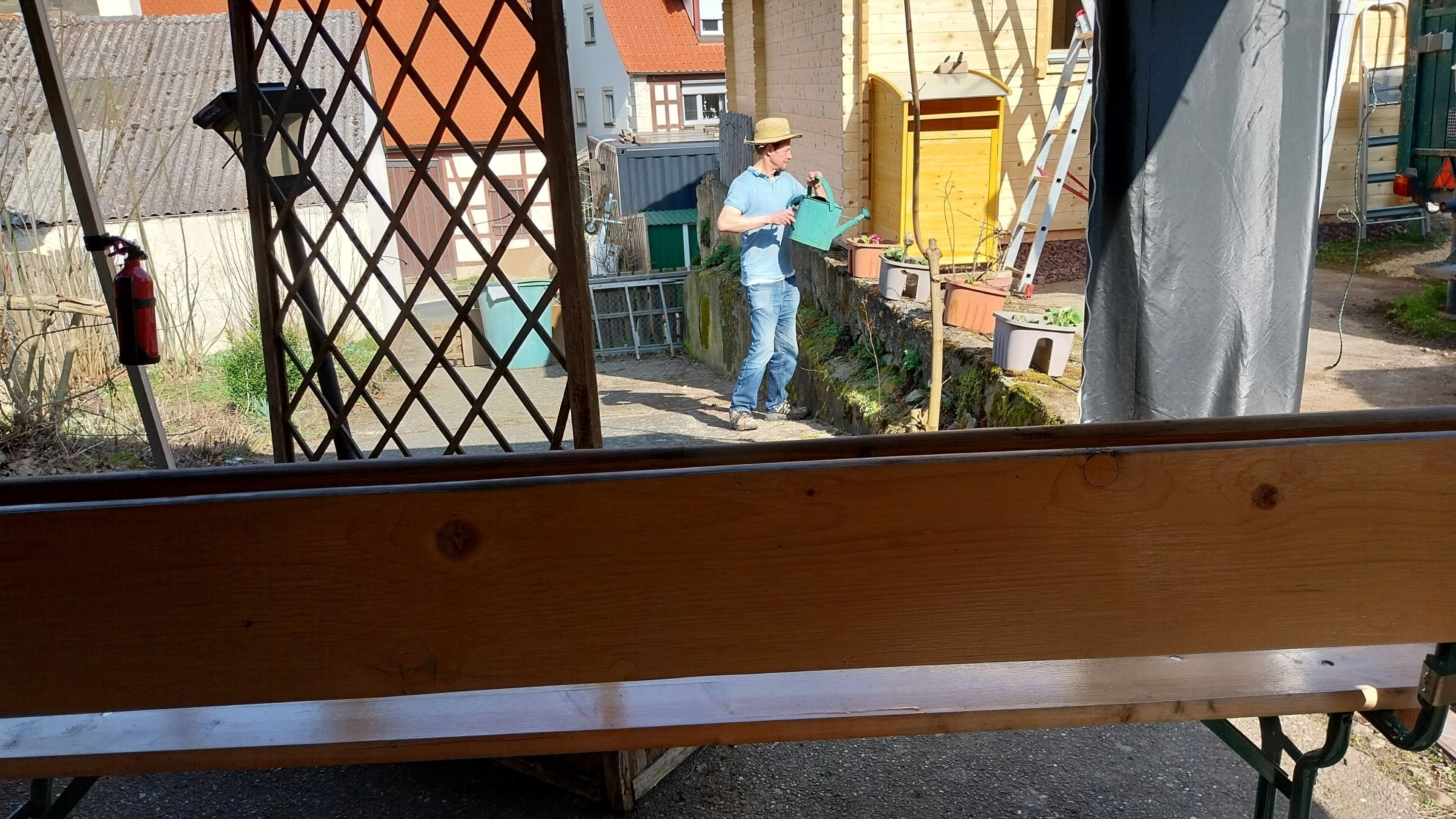 A wooden bench in the foreground with a view of a person watering plants in a garden area.