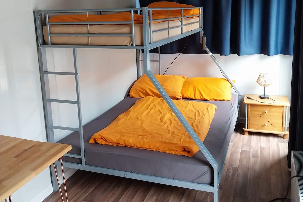 Una cama literera con ropa de cama naranja, un escritorio de madera, una mesita de noche con una lámpara y una ventana con cortinas azules.
