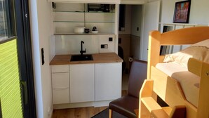 Un dormitorio pequeño con cama, una kitchenette con lavamanos y una silla.