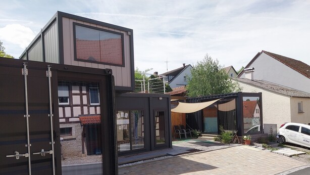 Un edificio moderno con un amplio ventanal, una zona de estar exterior cubierta y un automóvil estacionado.