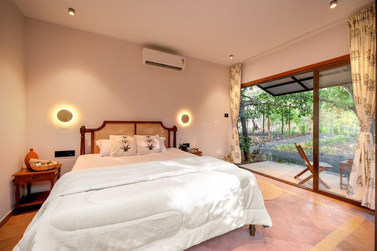 A bedroom with a large bed, a nightstand, a wall-mounted air conditioner, and a view of the outdoors through a sliding glass door.