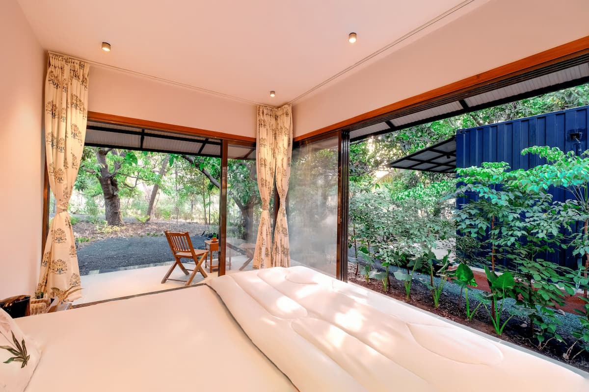 A room with a bed, a wooden chair, and large windows overlooking a garden.