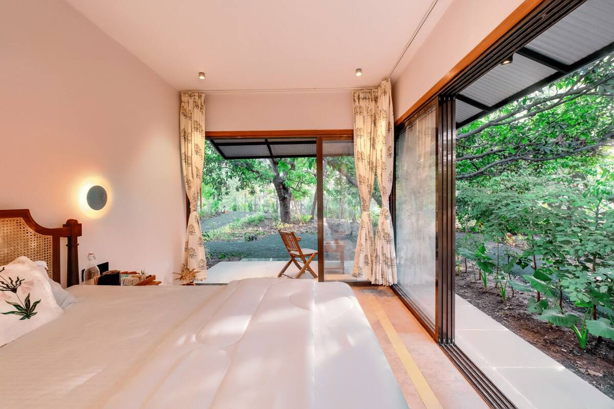 A bedroom with a large bed, a wooden headboard, a bedside table, and a chair. There is a window with curtains, and a view of greenery outside.