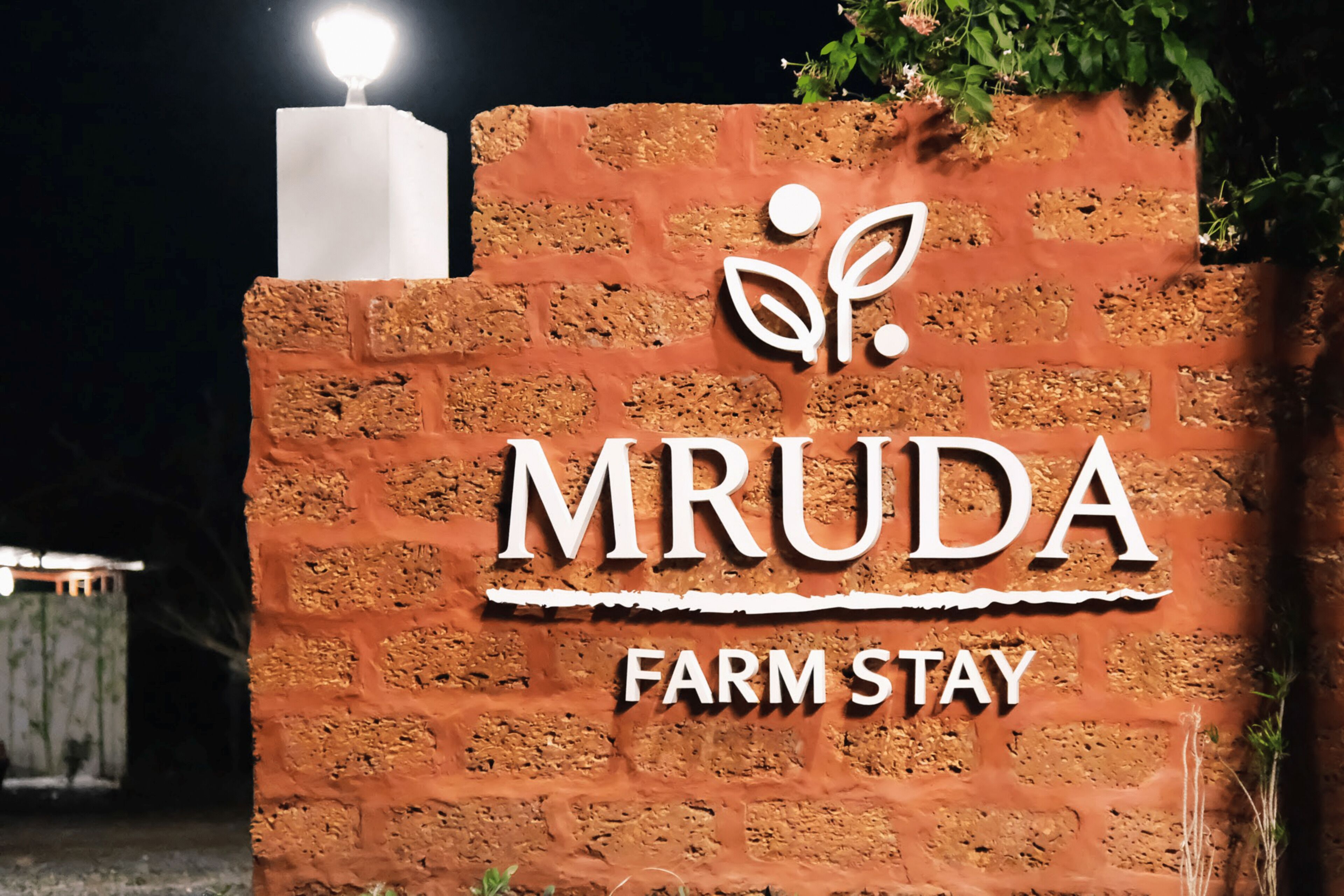 Un mur de briques sur lequel est apposée une enseigne indiquant « MRUDA FARM STAY », et une petite lumière blanche au-dessus.
