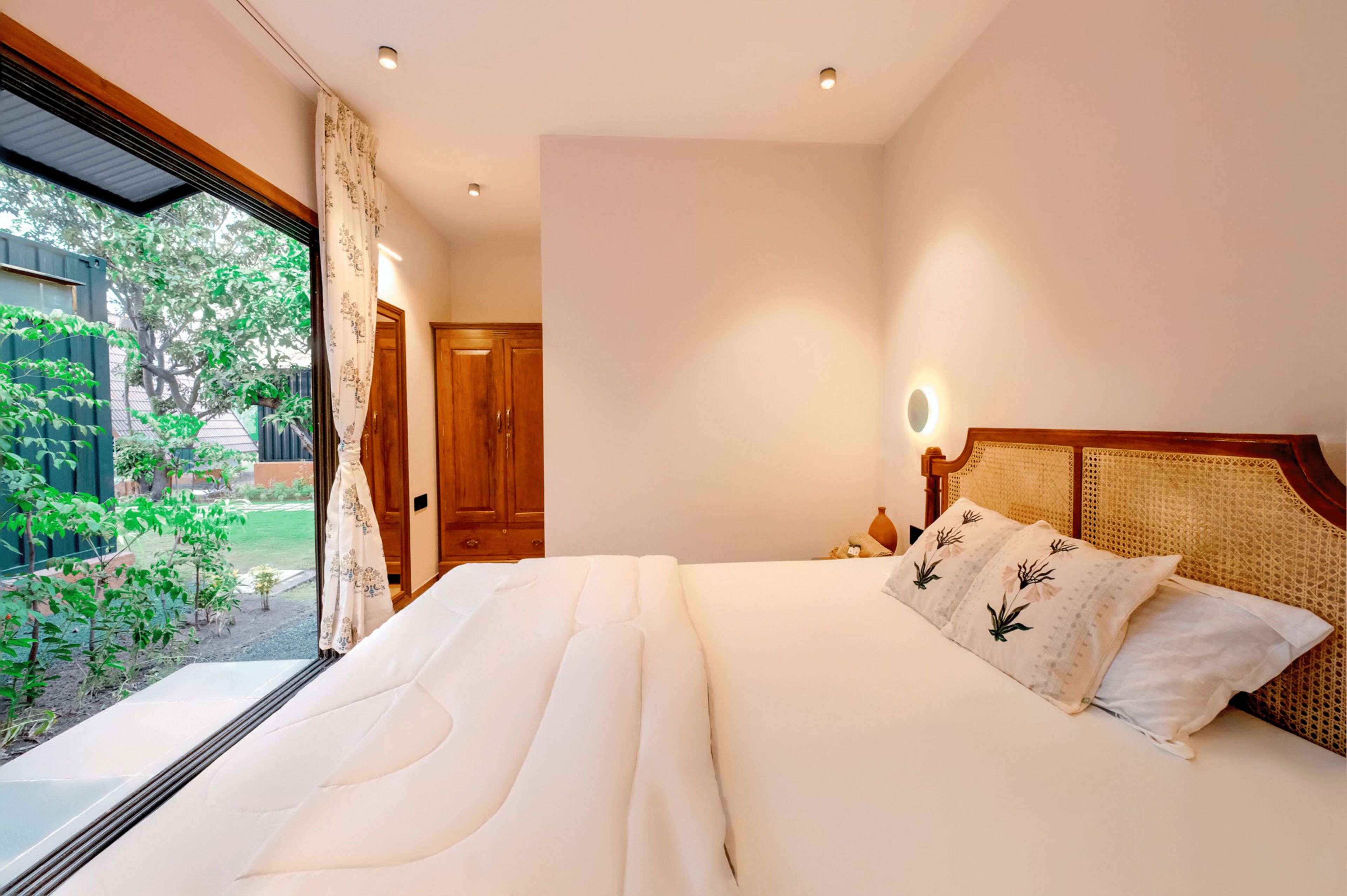 A bedroom with a large bed, a window with a view of greenery, a wooden door, and a bedside table with a lamp.