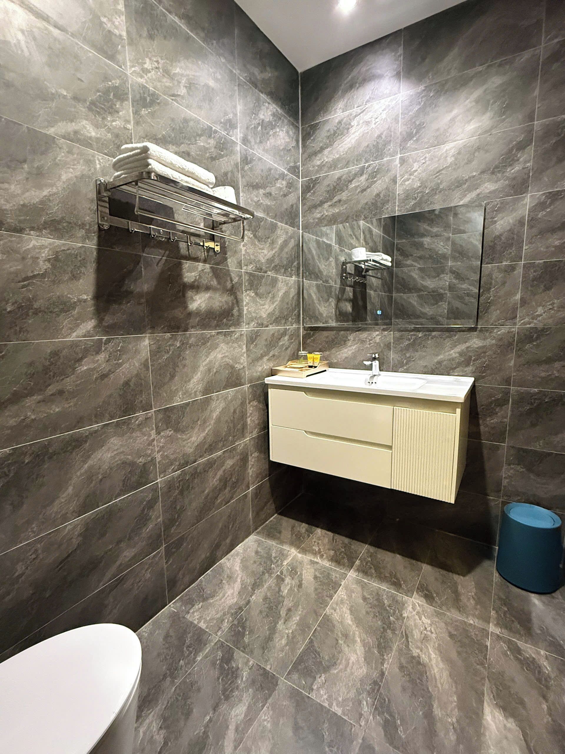 A modern bathroom with dark marble tiles, a white toilet, a beige vanity with a mirror, and a towel rack.