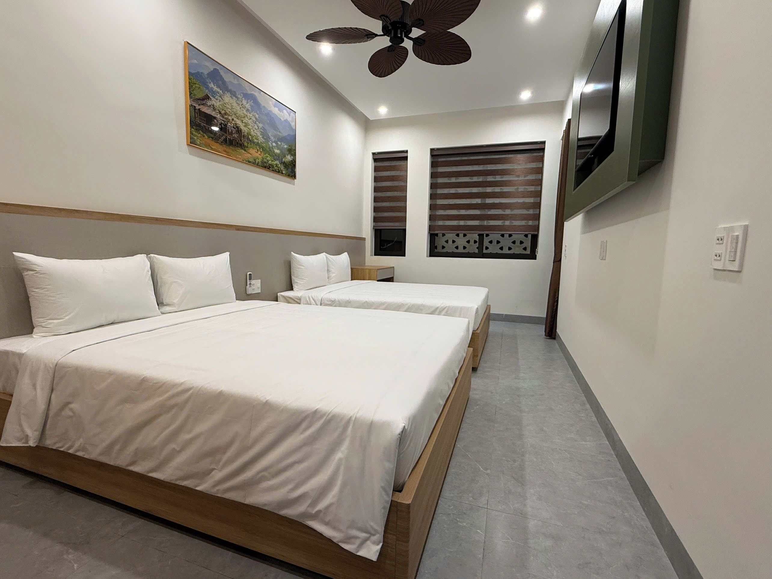 Habitación de hotel con dos camas, ventilador de techo, un cuadro y televisión en la pared.