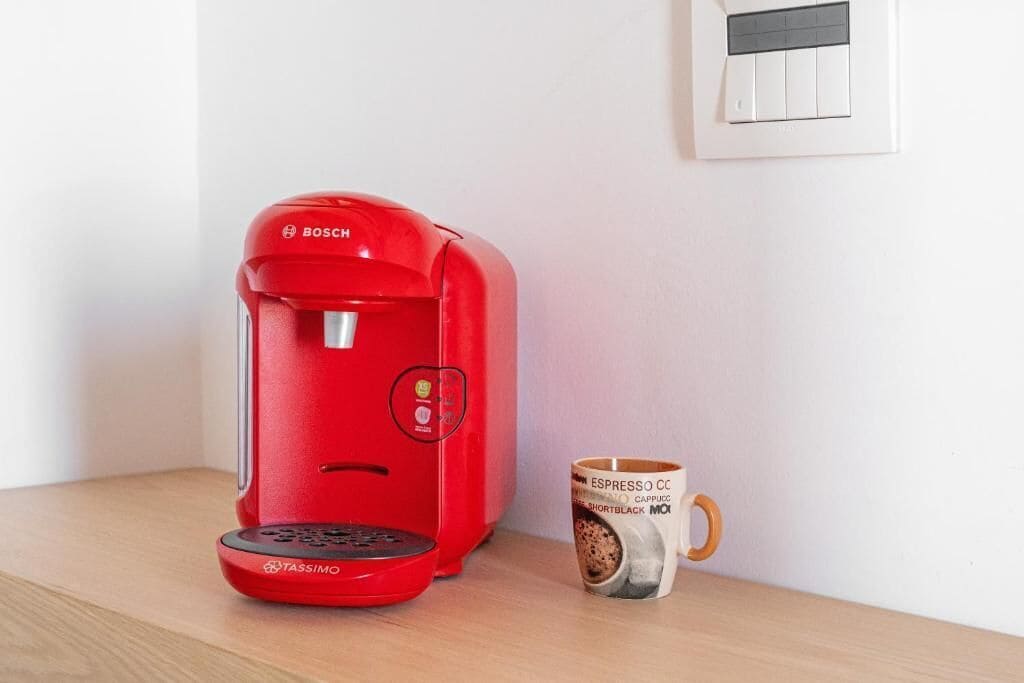 A red Tassimo coffee machine with a mug labeled "ESPRESSO CC CAPPUC SHORTBLACK MOK" on a wooden surface.