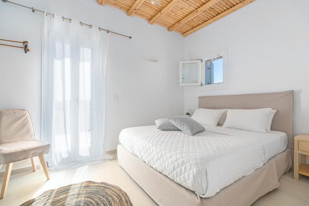 A bedroom with a bed, a chair, a bedside table, a mirror, and a window with curtains.