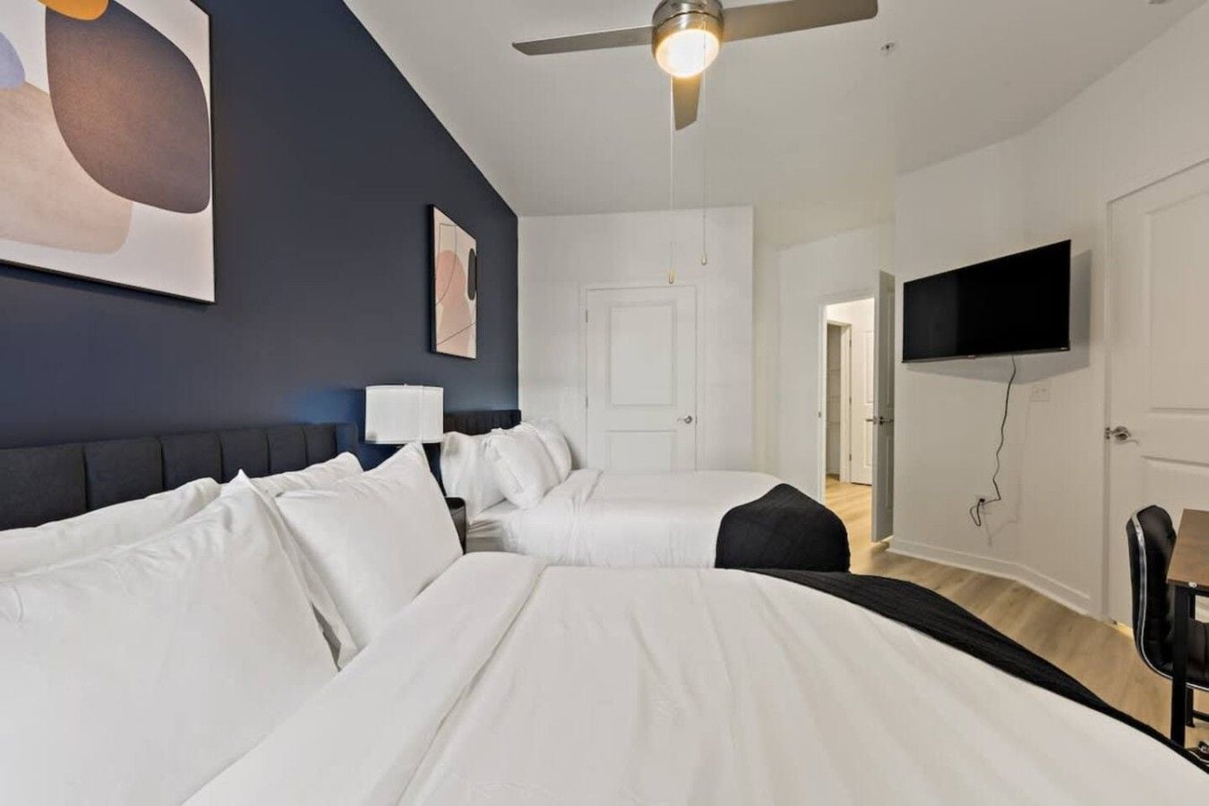 A hotel room with two beds, a ceiling fan, a flat-screen TV, and a door leading to another room.