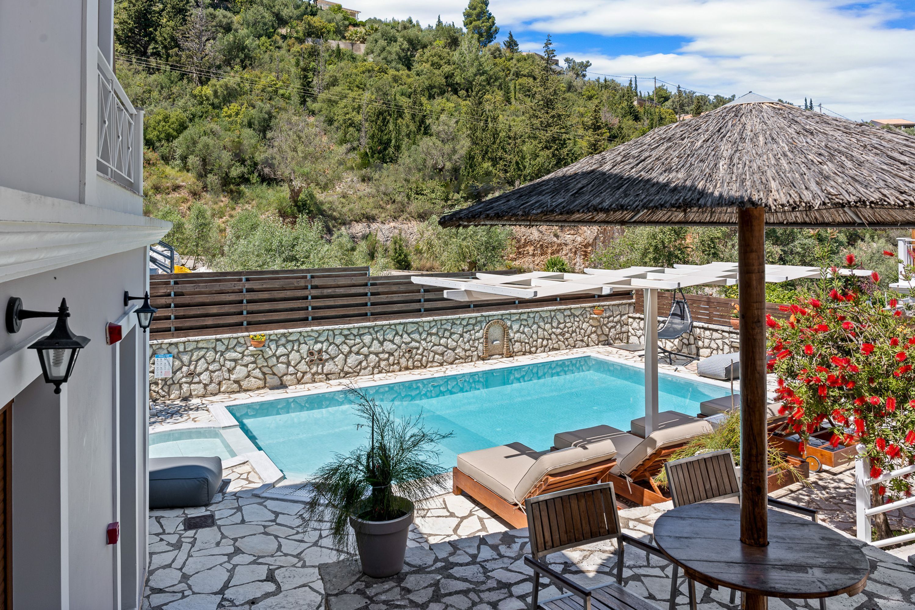 Un espace piscine doté d’un sol en pierre, de chaises longues, d’une table et d’un parasol en chaume.