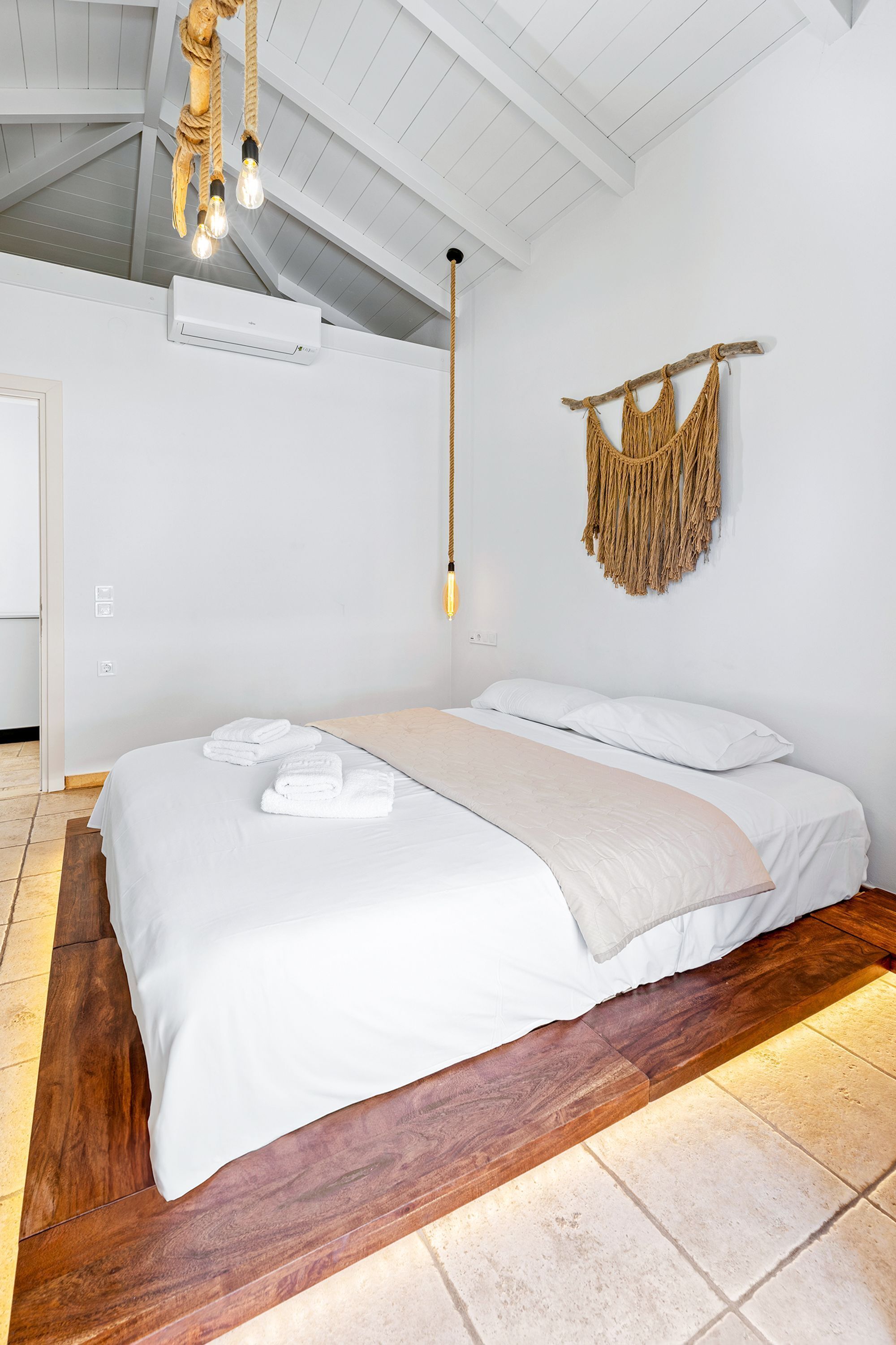A bedroom with a large bed, white bedding, a macrame wall hanging, and a hanging light fixture.