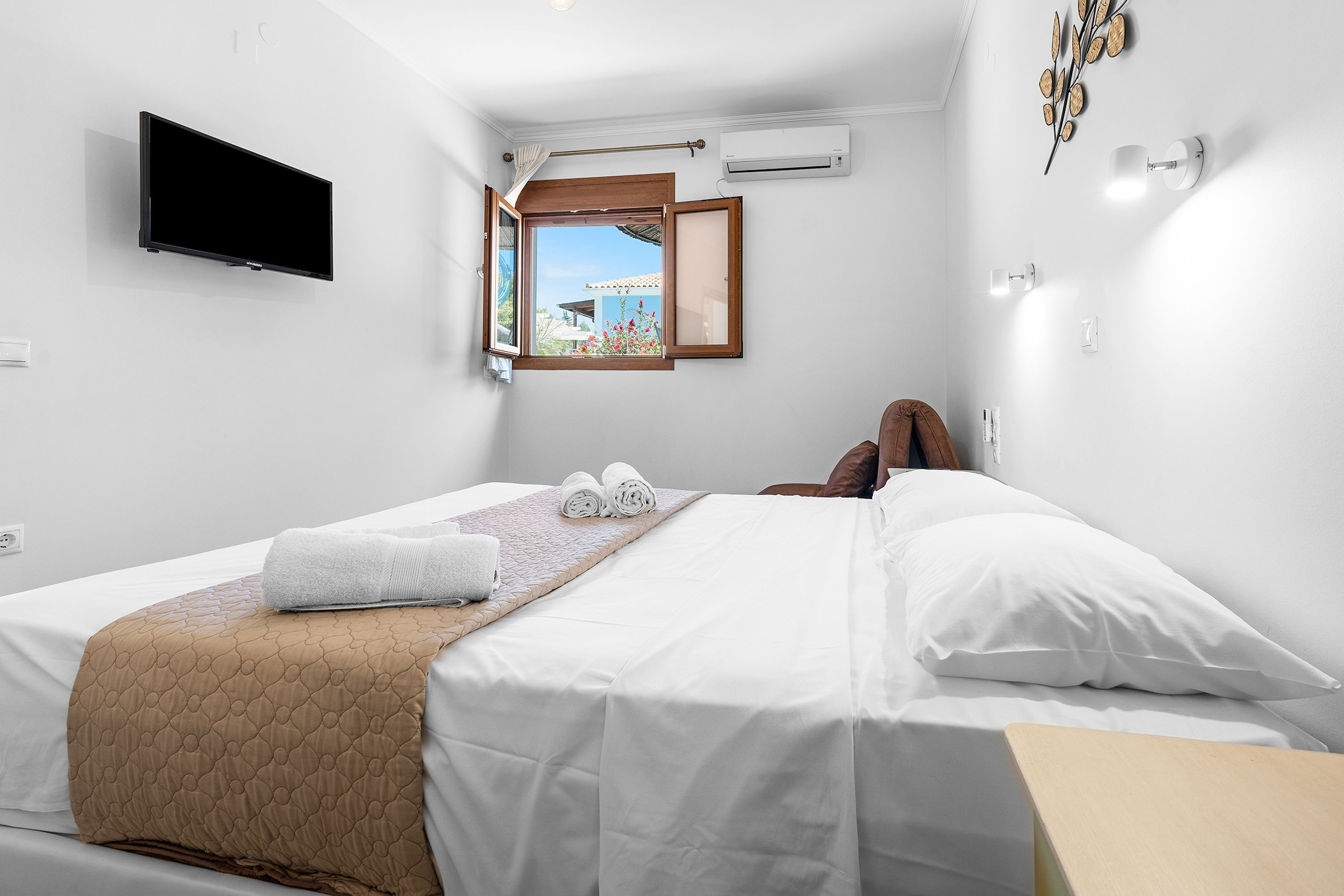 Ein Hotelzimmer mit einem Bett, einem an der Wand montierten Fernseher, einer Klimaanlage, einem Fenster mit geöffnetem Rollo und Blick ins Freie.