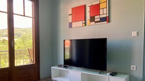 A living room with a flat-screen TV on a white cabinet, a window with a view of greenery, and abstract art on the wall. - Sea Breeze Apartment n 2 (Lefkada)