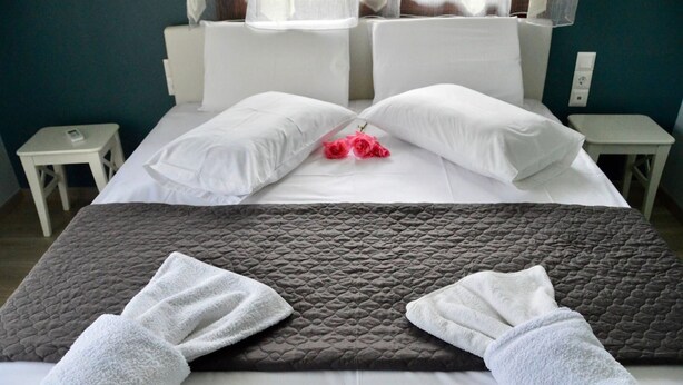 A neatly made bed with white pillows and a grey quilted blanket, two small bedside tables, and a pair of red roses on the pillows. - Sea Breeze Apartment n 2 (Lefkada)
