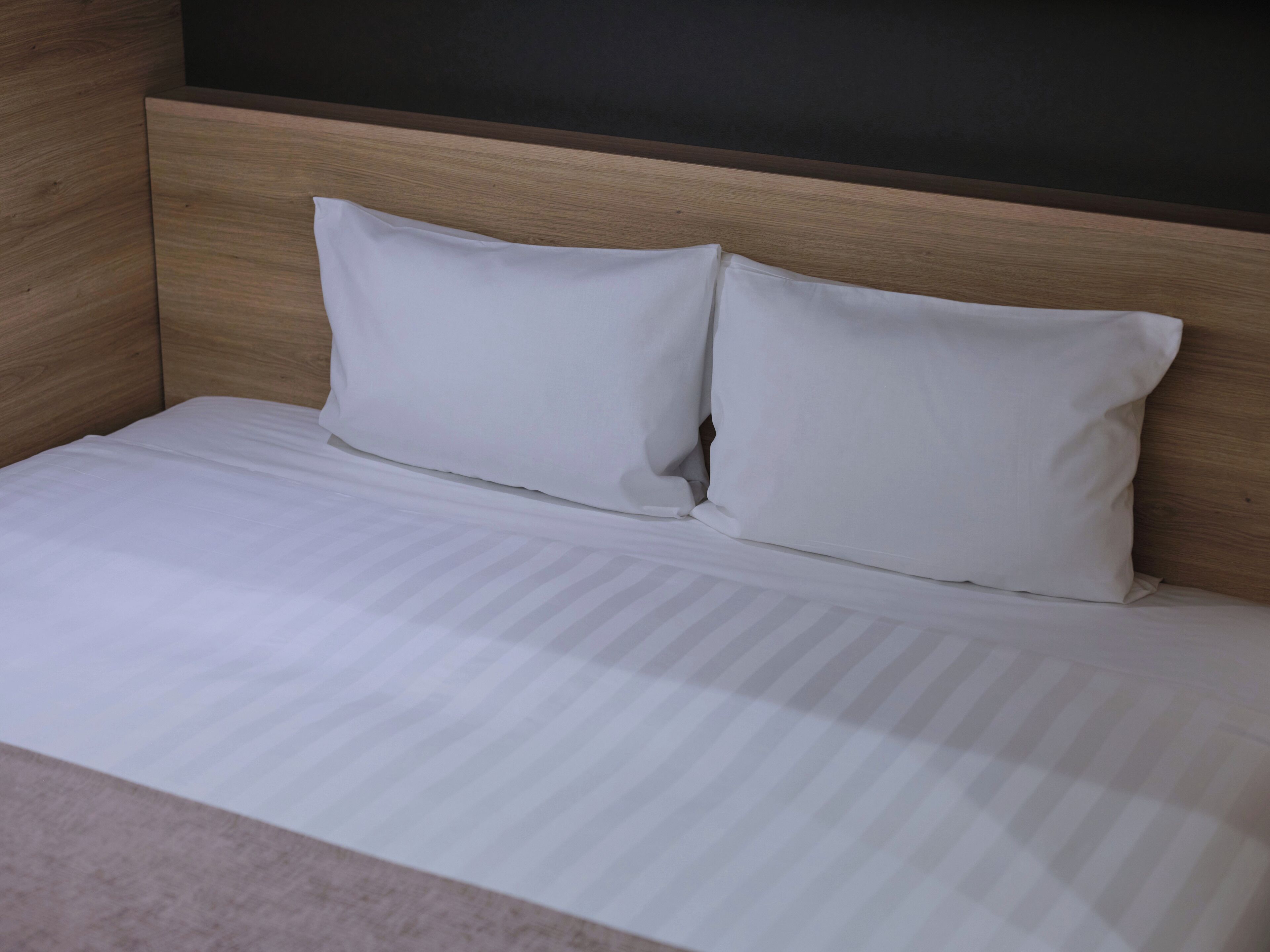 A bed with white bedding and two pillows, a wooden headboard, and a dark wall.
