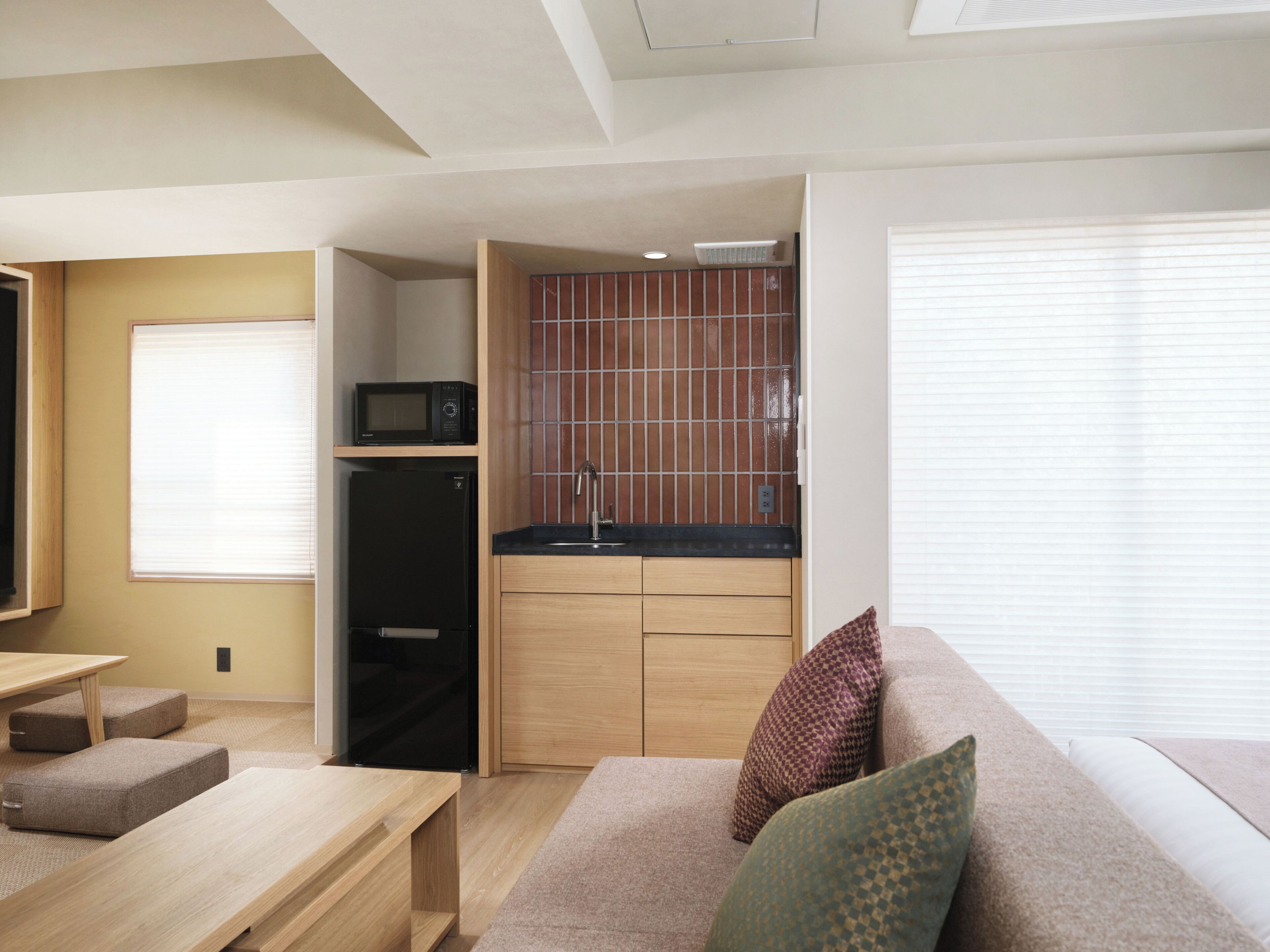 A compact living space with a kitchenette, a sofa with cushions, a small table, and a window with blinds.