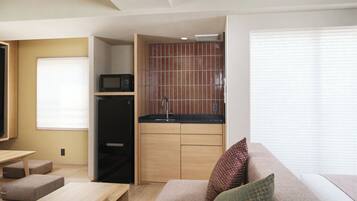 A compact living space with a kitchenette, a sofa with cushions, a small table, and a window with blinds.