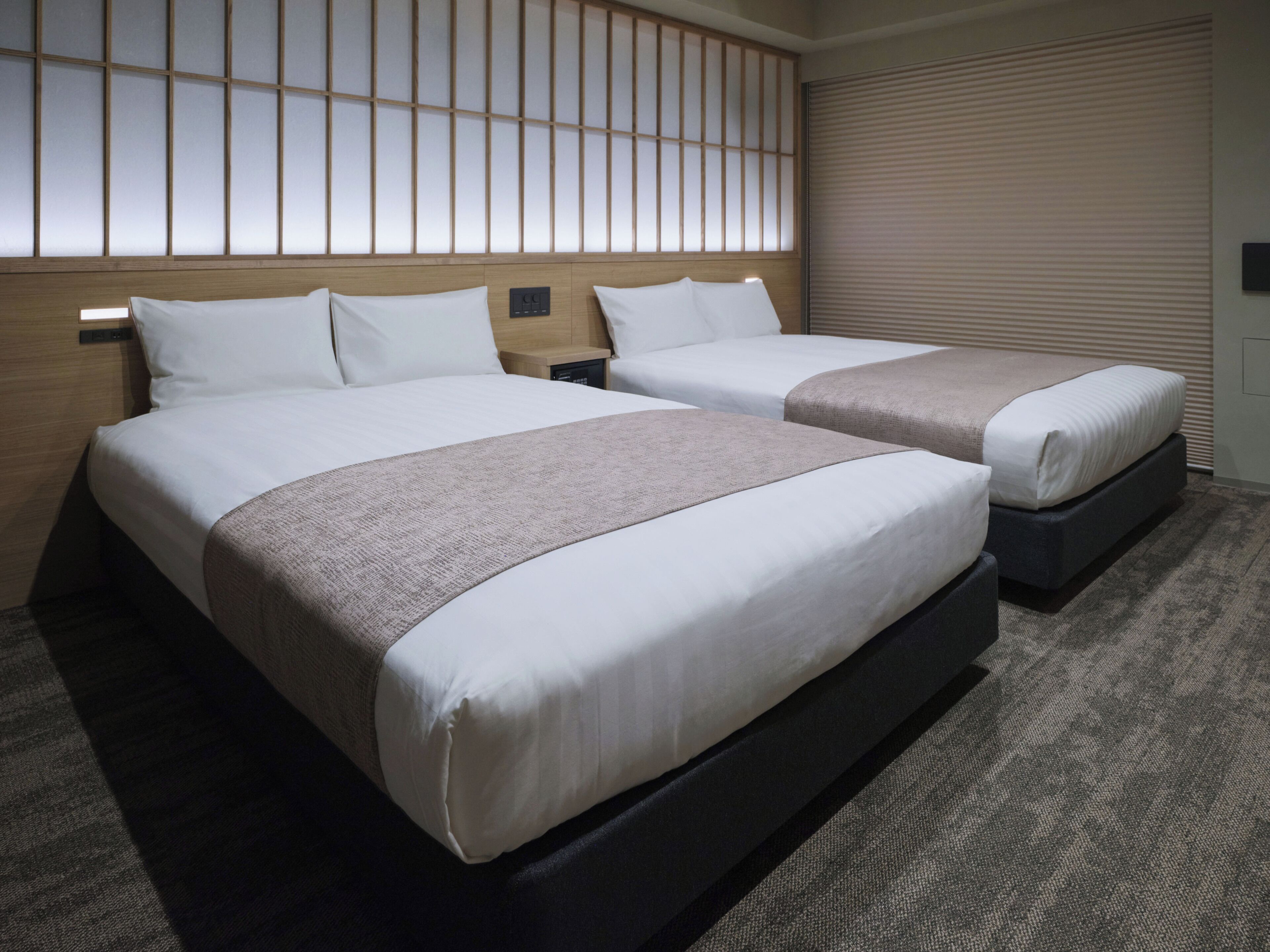 Quarto de hotel com duas camas, cabeceira de madeira, painel de controle e uma janela com persianas.
