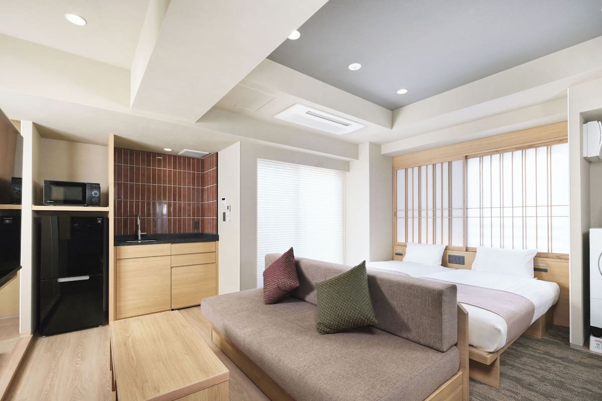 Habitación de hotel compacta con kitchenette, una cama con dos almohadas, una mesita de madera y una ventana con persianas.