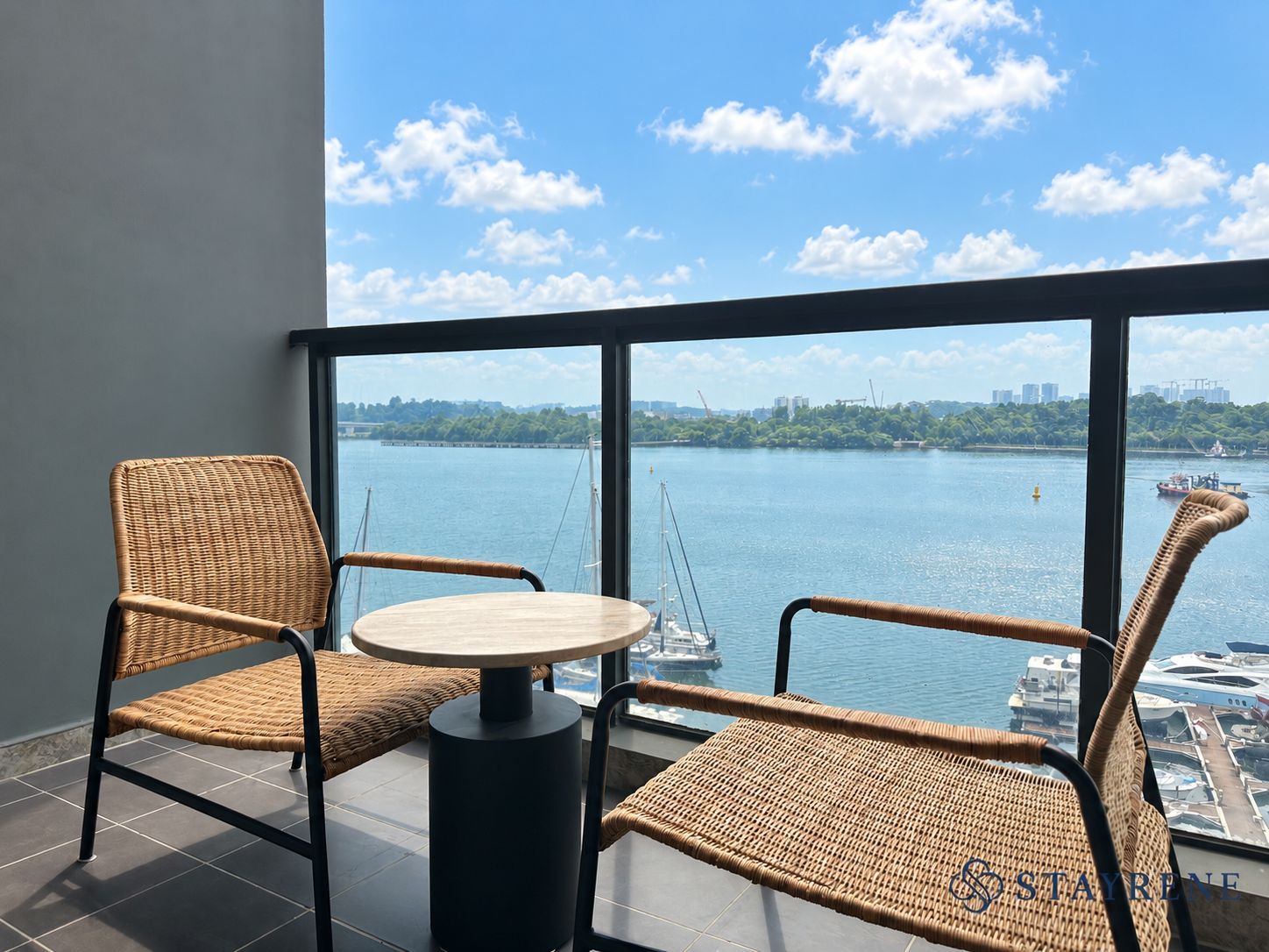 A balcony with wicker chairs and a round table, overlooking a body of water with boats and a distant cityscape.