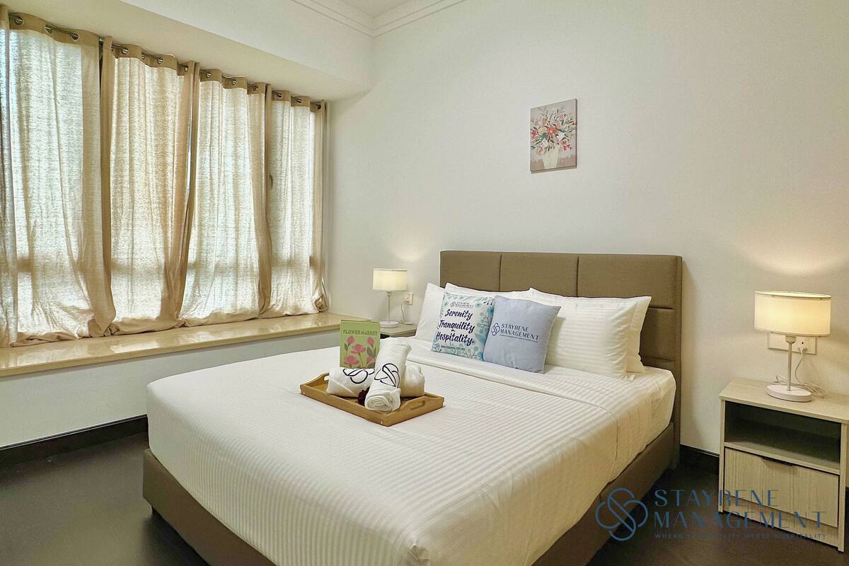 A hotel room with a large bed, a nightstand with a lamp, a window with curtains, and a small wall decoration.